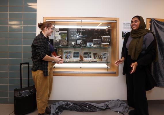 Wall display case containing photos and geological specimens presented on either side by two students. 