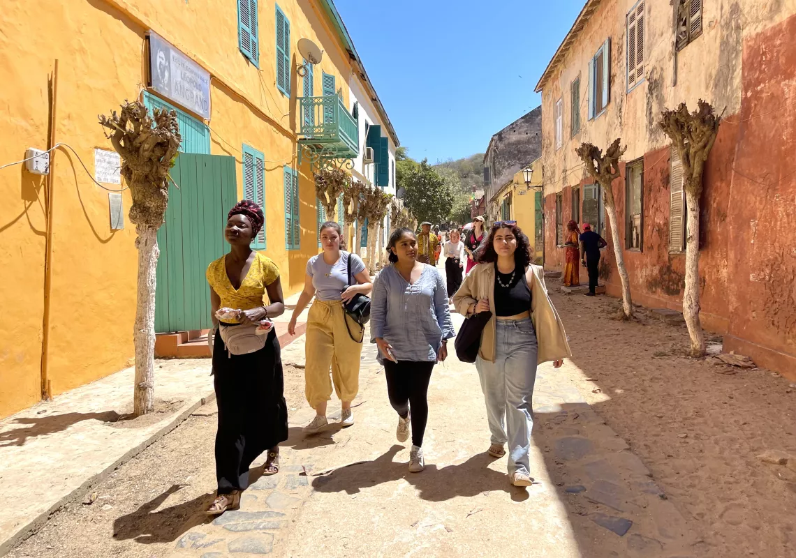 Four students walk down a dirt road with brightly colored two-storey buildings on either side.