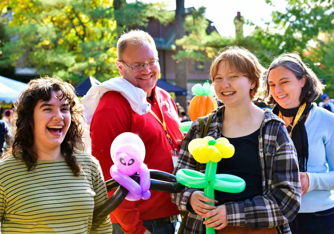A group of people, standing outside, hold balloon animals and laugh.