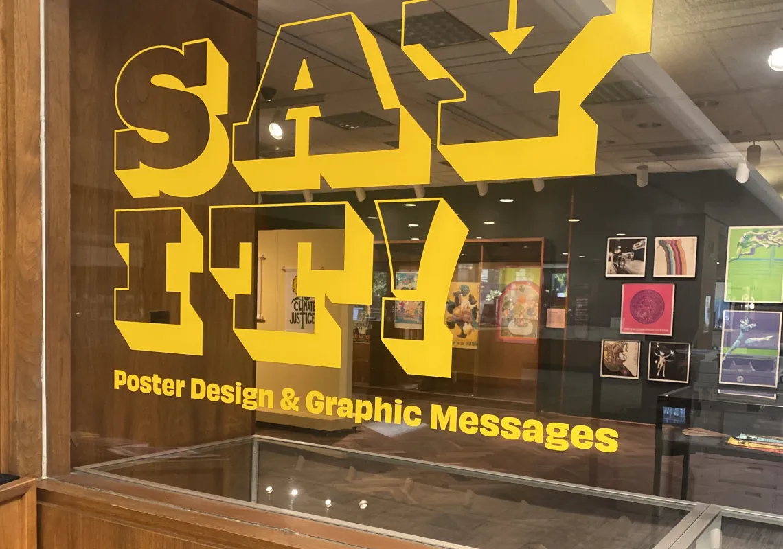 SAY IT! Poster Design & Graphic Messages is written in yellow vinyl on the window of the 1912 Gallery. Through the window you can see multicolored posters hanging on the wall.