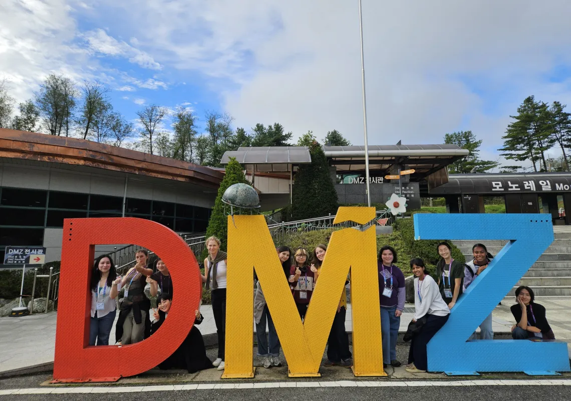 12 students behind a sculpture depicting the letters "D" "M" "Z" in primary colors.