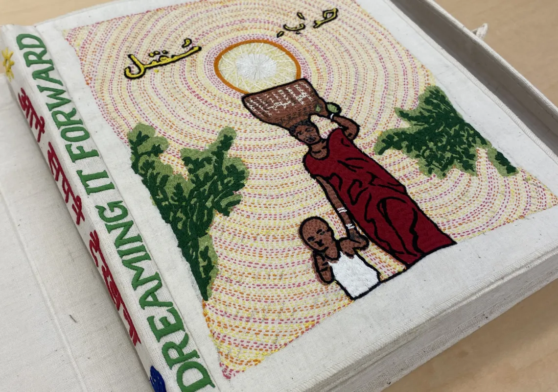 Cloth book with the words "Dreaming It Forward" stitched on the spine. The front image is a woman carrying a basket on her head while holding the hand of a child. There is Urdu text above her. 