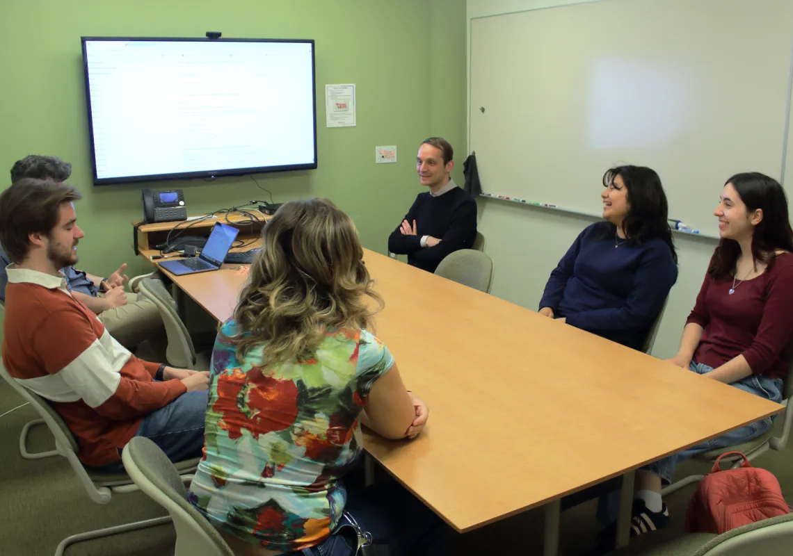 Faculty, students, and staff who worked Transnational Italian Studies projects talk around a long table. Project details are on a TV in the background.