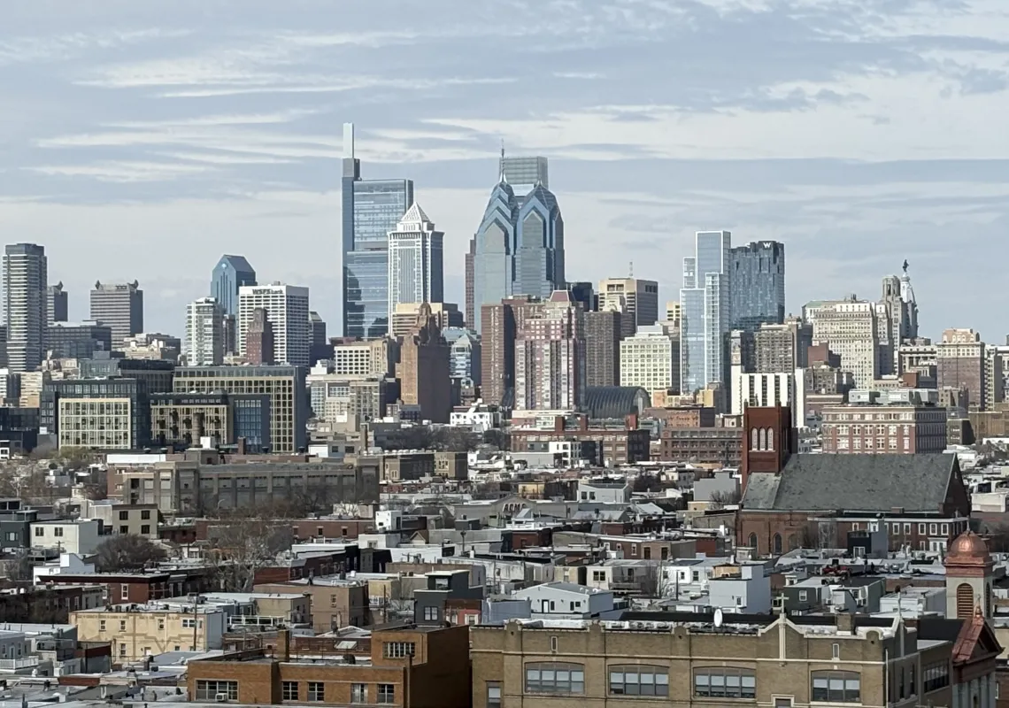 Image of view of Philly skyline from atop the Bok Building in South Philly, showing skyscrapers in the background, with low rise buildings and houses in the foreground, under a blue sky. Photo taken by Tanya Hoard.