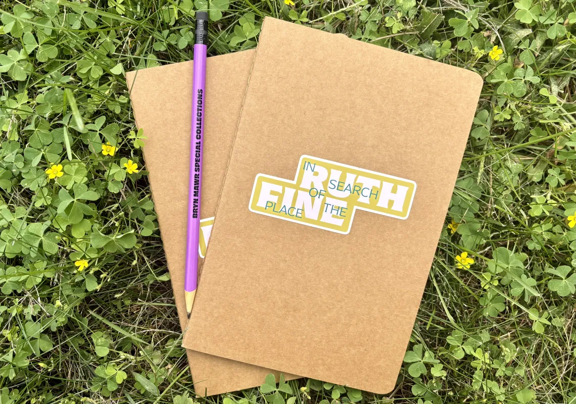 Two notebooks and a pencil sitting on the grass. The notebooks have a sticker on them that says "Ruth Fine: In Search of the Place." 