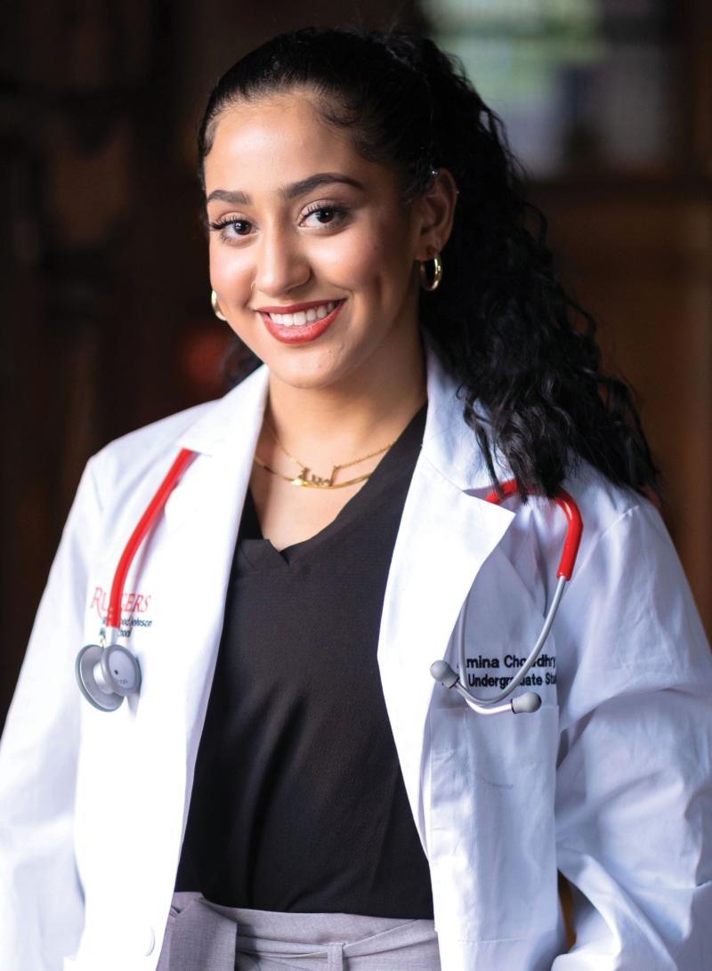 Amina Choudhry smiling, wearing a white lab coat and stethoscope