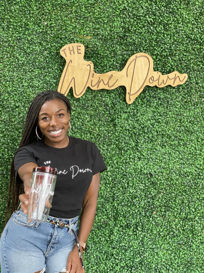 Chanelle Wilson standing and holding out a tumbler cup in front of a plant wall and a wooden sign that reads "The Wine Down"