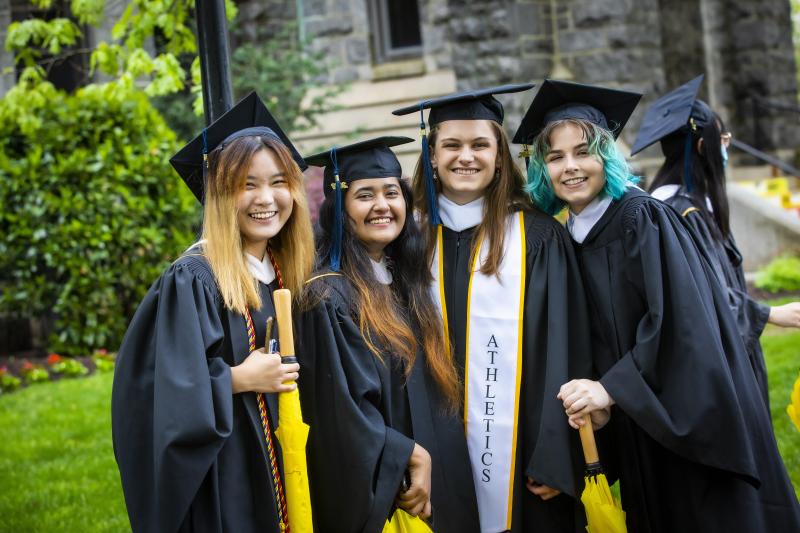 Photo of 4 members of the Class of 2022. They are smiling, wearing black graduate caps and gowns.