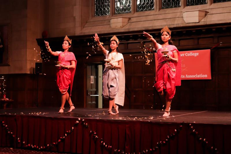 Three women wearing traditional Cambodian clothing stand on a stage sprinkling flower petals. 