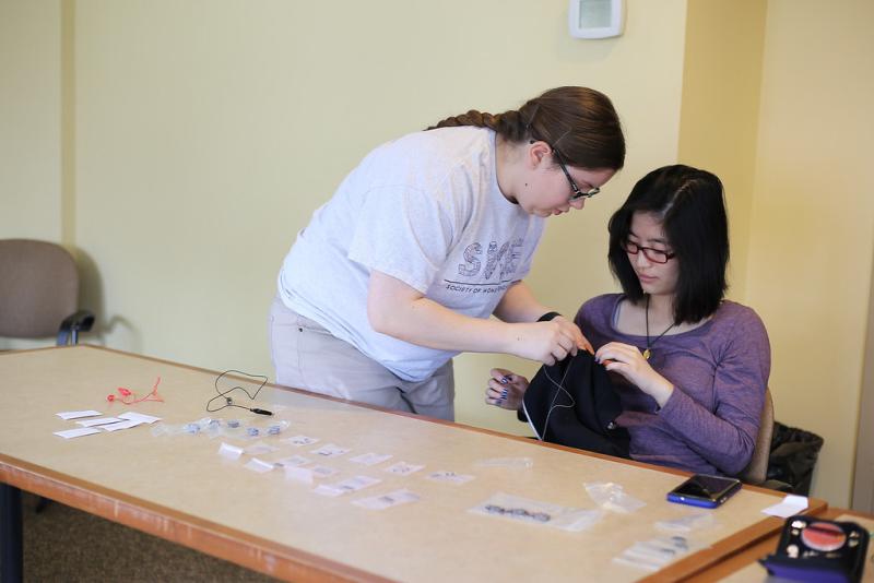 Two students sit at a table. One is sewing wires into fabric, the other is helping.