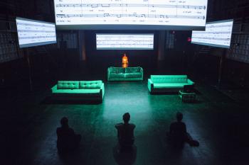 Three people sit on the floor across from three green couches on a dark stage. Four large projections of sheet music hang above the couches.