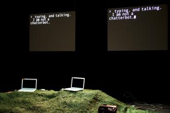 Two laptops perch on a grassy hill on a dark stage. Behind them, there are two black screens with the words "typing and talking, I am not a chatterbot" in white text.